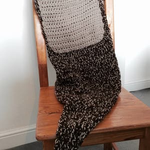 May include: A brown and cream crocheted blanket with a square patch in the center. The blanket is draped over a wooden chair.