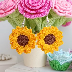 May include: Crocheted sunflower earrings with brown centres and silver hooks. The earrings are displayed against a background of crocheted pink roses.