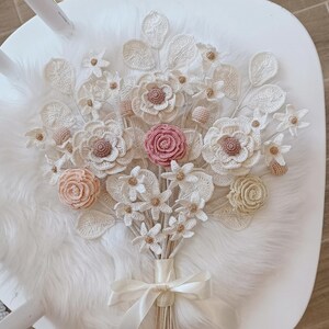 Puede incluir: Un ramo de flores de ganchillo hecho a mano con flores blancas, crema, rosas y beige. El ramo est&aacute; atado con una cinta de raso crema y descansa sobre una superficie blanca con una textura suave.