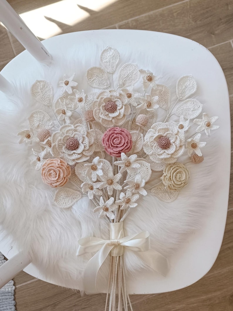 Puede incluir: Un ramo de flores de ganchillo hecho a mano con flores blancas, crema, rosas y beige. El ramo est&aacute; atado con una cinta de raso crema y descansa sobre una superficie blanca con una textura suave.