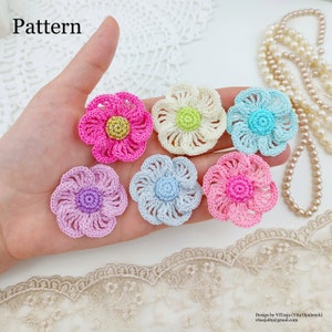 May include: Six crocheted flower embellishments in various colors, including pink, light blue, purple, cream, and light green. The flowers are arranged on a hand, with a lace background.