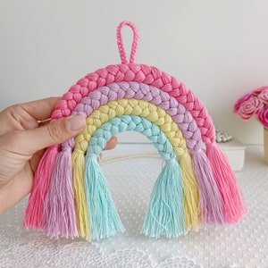 Puede incluir: Un tapiz de macramé de arcoíris con un diseño de cuerda trenzada y borlas en rosa, morado, amarillo y azul.