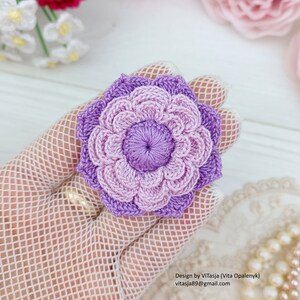 Puede incluir: Una flor de ganchillo con un efecto degradado de color morado y morado claro. La flor tiene un di&aacute;metro de aproximadamente 7,5 cm.