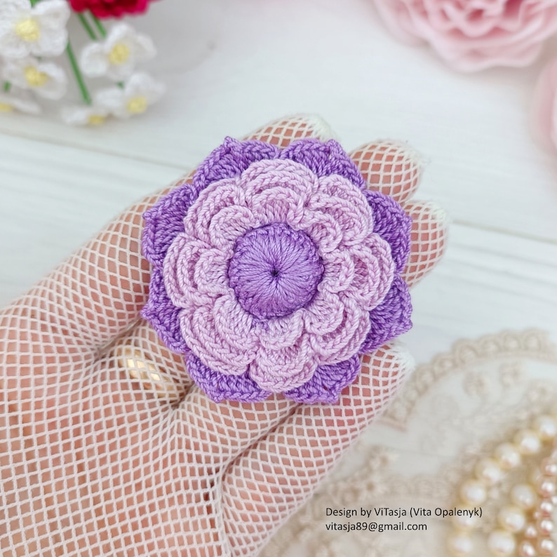 Puede incluir: Una flor de ganchillo con un efecto degradado de color morado y morado claro. La flor tiene un di&aacute;metro de aproximadamente 7,5 cm.