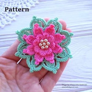 Könnte beinhalten: Eine gehäkelte Blume mit einem rosa Zentrum und grünen Blättern. Die Blume ist mit weißen Perlen verziert.