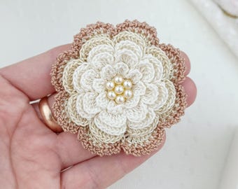 May include: A multi-layered crochet flower brooch featuring cream and beige yarn petals with a cluster of six small, round, off-white faux pearls at the center. The outer edge of the flower is finished with a darker beige yarn trim.