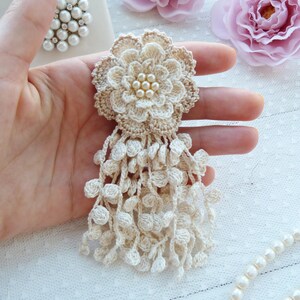 Puede incluir: Un broche de flor de ganchillo en color beige con un dise&ntilde;o de p&eacute;talos superpuestos, un centro adornado con un grupo de peque&ntilde;as perlas blancas y un largo fleco colgante formado por numerosas esferas peque&ntilde;as de ganchillo.