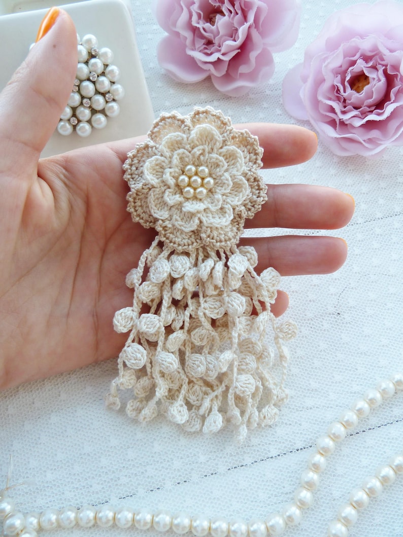 Puede incluir: Un broche de flor de ganchillo en color beige con un dise&ntilde;o de p&eacute;talos superpuestos, un centro adornado con un grupo de peque&ntilde;as perlas blancas y un largo fleco colgante formado por numerosas esferas peque&ntilde;as de ganchillo.