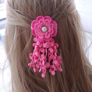 Puede incluir: Un accesorio para el cabello tejido a ganchillo en color rosa con un dise&ntilde;o de flor de varias capas, un bot&oacute;n blanco central y m&uacute;ltiples hebras colgantes tejidas con peque&ntilde;os detalles circulares. El texto ViTasja aparece arriba a la derecha.