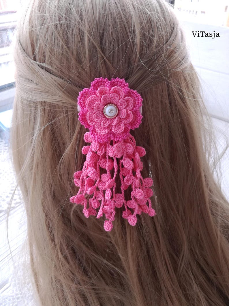 Puede incluir: Un accesorio para el cabello tejido a ganchillo en color rosa con un dise&ntilde;o de flor de varias capas, un bot&oacute;n blanco central y m&uacute;ltiples hebras colgantes tejidas con peque&ntilde;os detalles circulares. El texto ViTasja aparece arriba a la derecha.