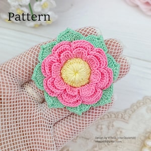 Puede incluir: Una flor de crochet con p&eacute;talos rosas y un centro amarillo, rodeada de hojas verdes. La flor est&aacute; sobre un fondo blanco.