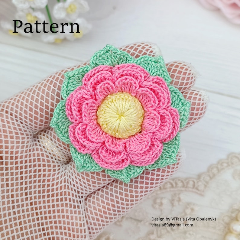 Puede incluir: Una flor de crochet con p&eacute;talos rosas y un centro amarillo, rodeada de hojas verdes. La flor est&aacute; sobre un fondo blanco.