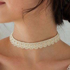 May include: Cream crochet choker necklace with a scalloped edge and small pearl accents. The delicate design is visible against the skin, showcasing the intricate craftsmanship. This handmade necklace is a charming accessory.