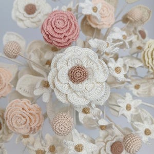 Puede incluir: Primer plano de un arreglo floral artesanal con flores de ganchillo de varios tama&ntilde;os y estilos. La paleta de colores incluye blanco, crema, rosa rubor y beige. El arreglo incluye rosas, margaritas y otros tipos de flores, con hojas y tallos.