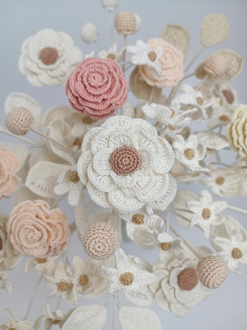 Puede incluir: Primer plano de un arreglo floral artesanal con flores de ganchillo de varios tama&ntilde;os y estilos. La paleta de colores incluye blanco, crema, rosa rubor y beige. El arreglo incluye rosas, margaritas y otros tipos de flores, con hojas y tallos.