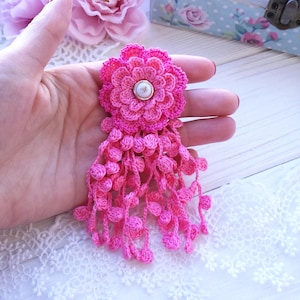 Puede incluir: Un broche de flor de ganchillo rosa brillante con un dise&ntilde;o de p&eacute;talos en capas, un bot&oacute;n blanco central como acento y m&uacute;ltiples hilos colgantes de peque&ntilde;as esferas de ganchillo que cuelgan de la base.