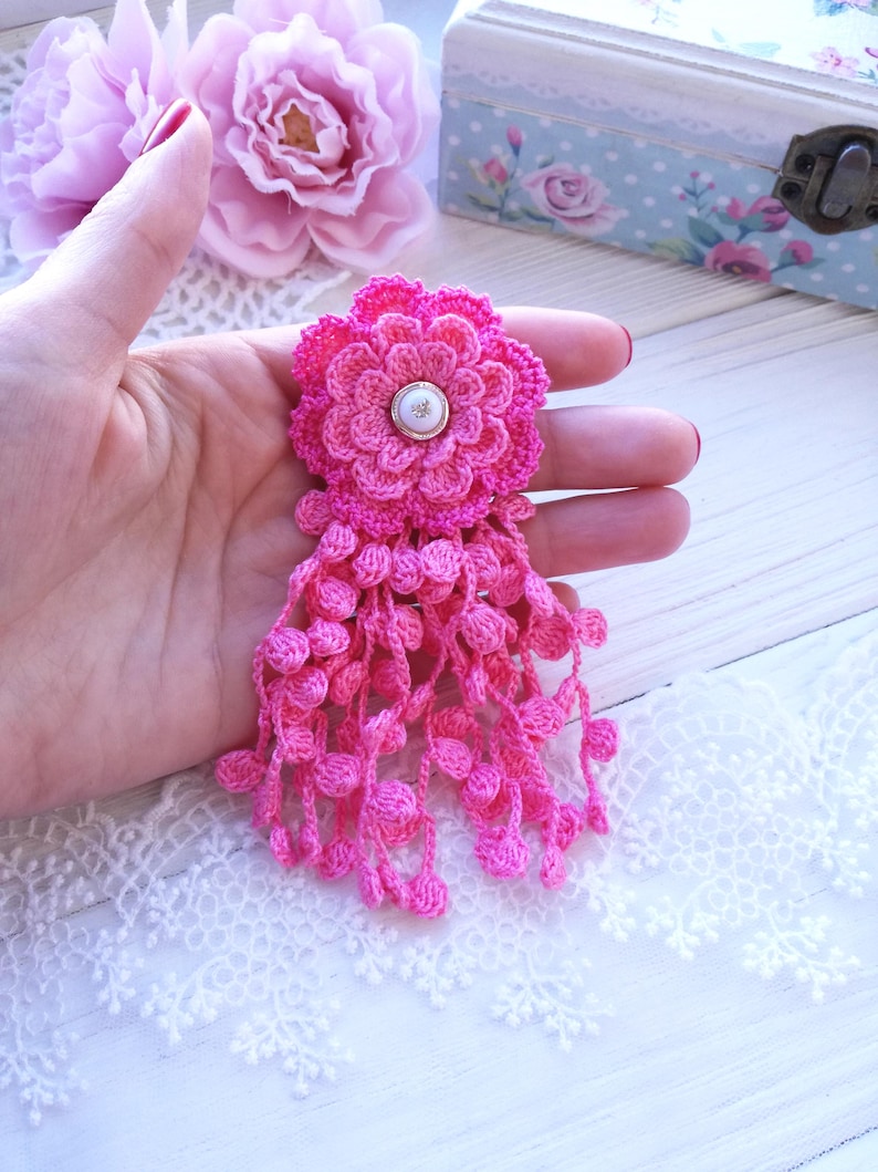 Puede incluir: Un broche de flor de ganchillo rosa brillante con un dise&ntilde;o de p&eacute;talos en capas, un bot&oacute;n blanco central como acento y m&uacute;ltiples hilos colgantes de peque&ntilde;as esferas de ganchillo que cuelgan de la base.