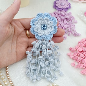 Puede incluir: Un broche de flor de ganchillo azul claro con un dise&ntilde;o de p&eacute;talos superpuestos, un centro de peque&ntilde;as cuentas azules y un largo fleco colgante hecho de delicados hilos de ganchillo texturizados que cuelgan de la base.