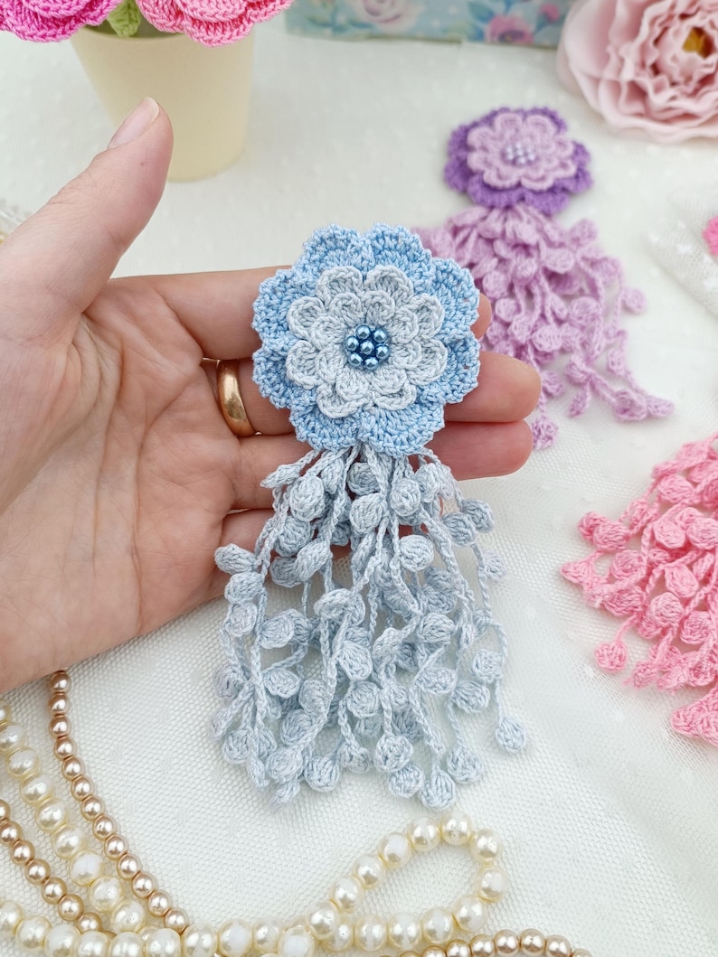 Puede incluir: Un broche de flor de ganchillo azul claro con un dise&ntilde;o de p&eacute;talos superpuestos, un centro de peque&ntilde;as cuentas azules y un largo fleco colgante hecho de delicados hilos de ganchillo texturizados que cuelgan de la base.