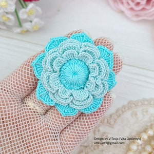 Puede incluir: Un broche de flor tejido a crochet en tonos de azul claro y oscuro. La flor tiene un bot&oacute;n central y est&aacute; hecha con un delicado patr&oacute;n de puntadas.