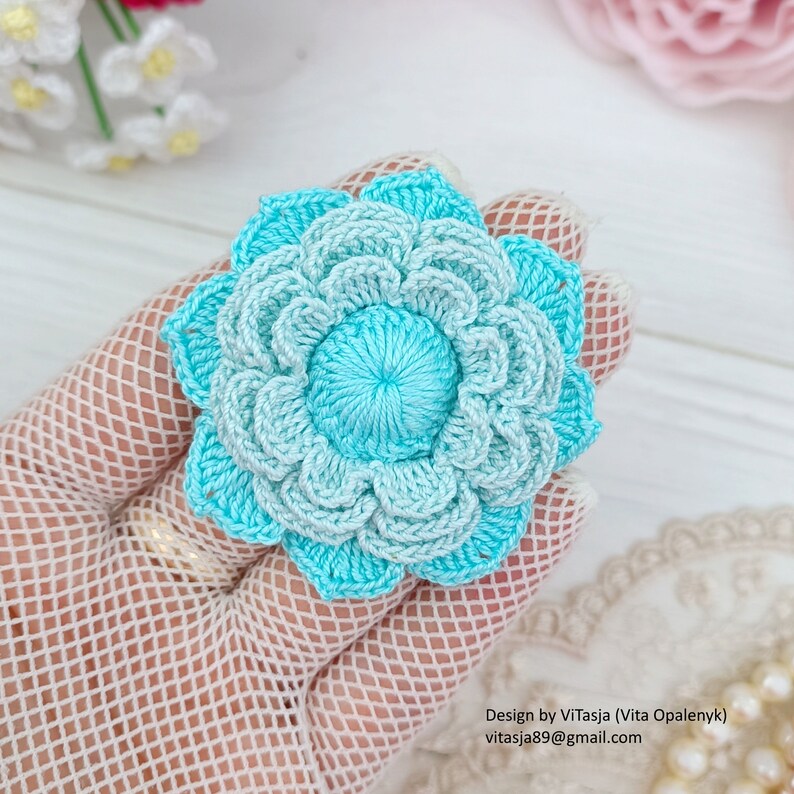 Puede incluir: Un broche de flor tejido a crochet en tonos de azul claro y oscuro. La flor tiene un bot&oacute;n central y est&aacute; hecha con un delicado patr&oacute;n de puntadas.
