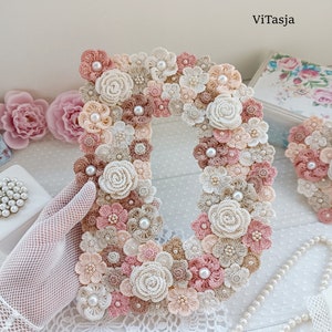 May include: A crocheted letter D made of pink, beige and white flowers with pearl accents. The letter is displayed on a white lace tablecloth.