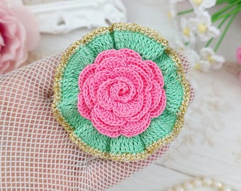 May include: A circular crochet flower brooch featuring a vibrant pink rose center surrounded by a ruffled mint green border, all finished with a delicate metallic gold thread trim along the outer edge of the circular piece.