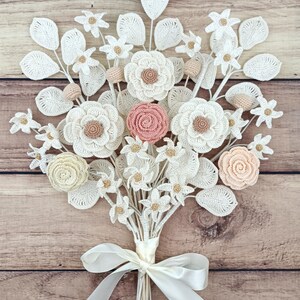 Puede incluir: Un ramo artesanal de flores de ganchillo en tonos blanco, crema y melocot&oacute;n. El ramo presenta varios tipos de flores, incluyendo rosas y margaritas, con detalles de hojas. Atado con una cinta de raso blanca, descansa sobre un fondo de madera.