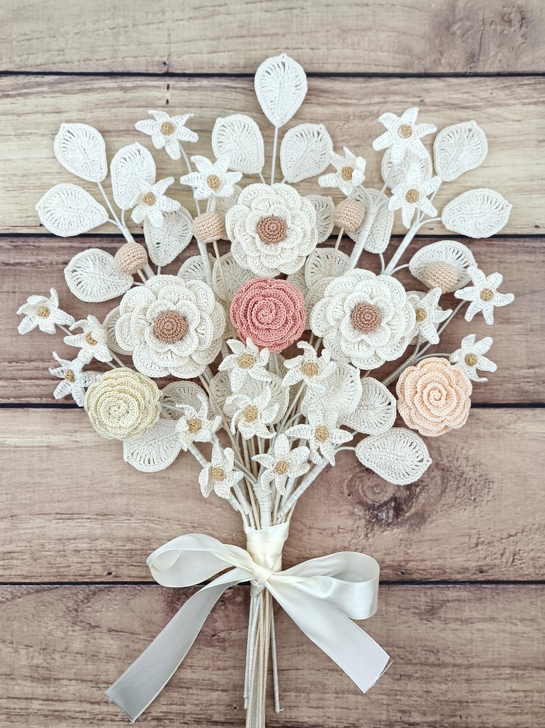 Puede incluir: Un ramo artesanal de flores de ganchillo en tonos blanco, crema y melocot&oacute;n. El ramo presenta varios tipos de flores, incluyendo rosas y margaritas, con detalles de hojas. Atado con una cinta de raso blanca, descansa sobre un fondo de madera.