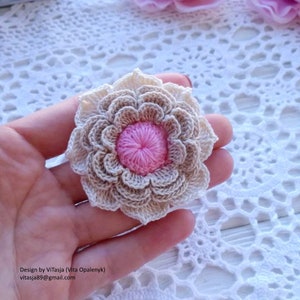Puede incluir: Una flor de ganchillo con un centro rosa y p&eacute;talos beige. La flor se sostiene en una mano sobre un fondo de ganchillo blanco.