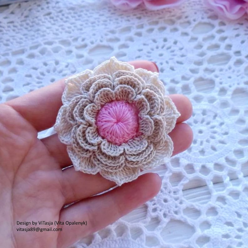 Puede incluir: Una flor de ganchillo con un centro rosa y p&eacute;talos beige. La flor se sostiene en una mano sobre un fondo de ganchillo blanco.