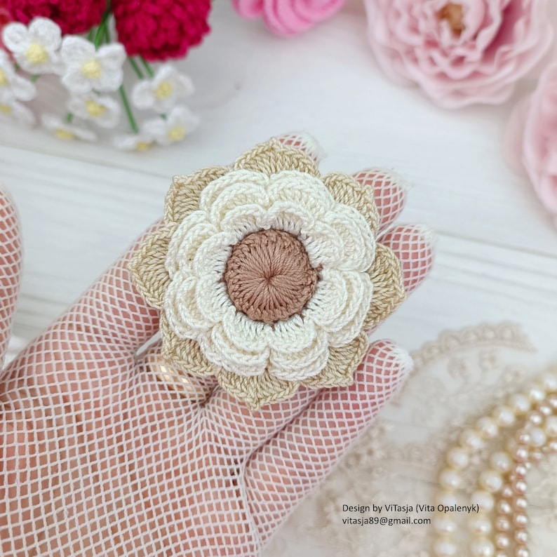 Puede incluir: Un broche de flor tejido a crochet con un dise&ntilde;o en capas beige y blanco. El centro de la flor es un c&iacute;rculo marr&oacute;n m&aacute;s peque&ntilde;o.
