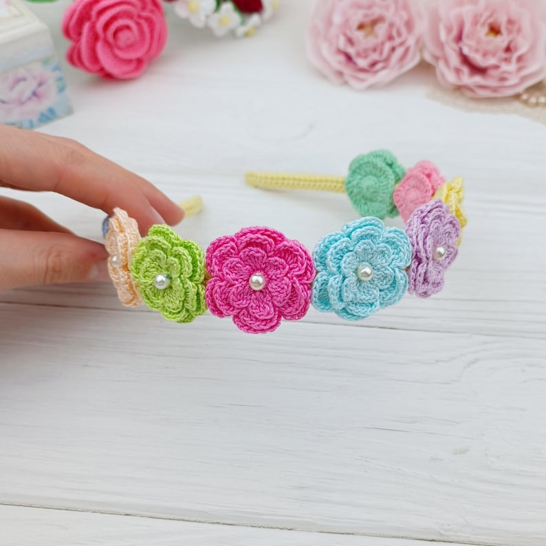 May include: A handmade crochet headband featuring colorful flowers in shades of pink, green, blue, and purple. Each flower has a small pearl in the center. The headband is yellow and the flowers are attached to it.