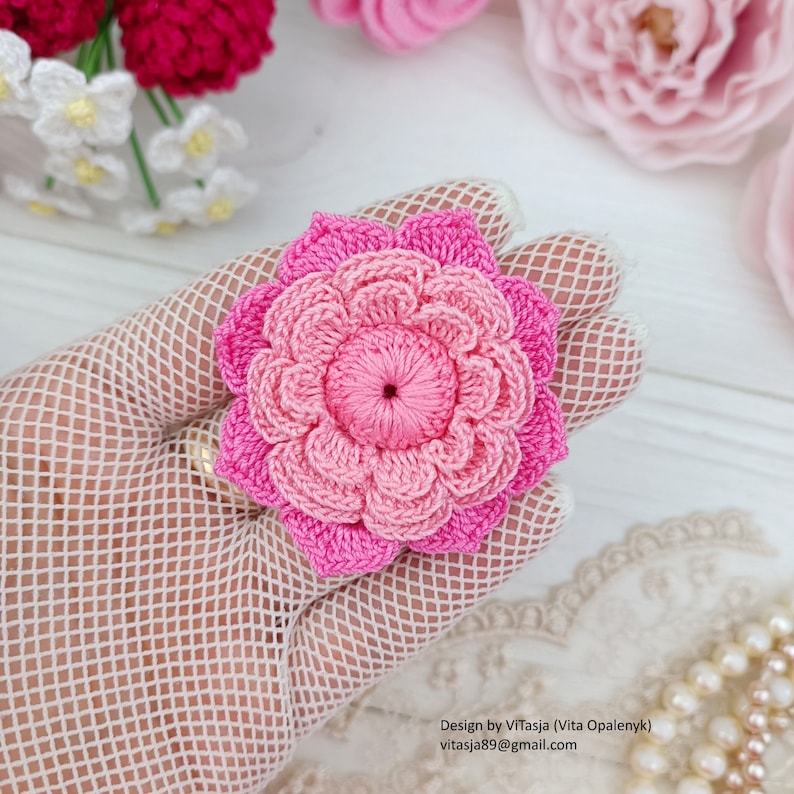 Puede incluir: Una flor de crochet rosa con un dise&ntilde;o en capas. La flor est&aacute; hecha con dos tonos de hilo rosa y tiene un peque&ntilde;o centro.