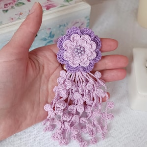 Puede incluir: Un broche de flor de ganchillo en tonos violeta claro y oscuro, con un dise&ntilde;o de p&eacute;talos en capas y un grupo de peque&ntilde;as cuentas blancas en el centro. La flor est&aacute; unida a un largo fleco colgante hecho de esferas de ganchillo violeta claro.