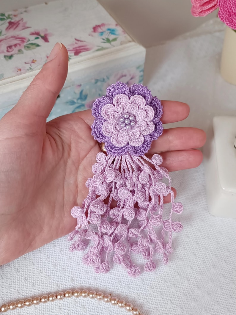 Puede incluir: Un broche de flor de ganchillo en tonos violeta claro y oscuro, con un dise&ntilde;o de p&eacute;talos en capas y un grupo de peque&ntilde;as cuentas blancas en el centro. La flor est&aacute; unida a un largo fleco colgante hecho de esferas de ganchillo violeta claro.
