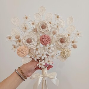 Puede incluir: Un ramo de flores de ganchillo hecho a mano con flores crema, melocot&oacute;n y rosa. El ramo presenta diferentes tipos de flores, detalles de p&eacute;talos y un tallo atado con una cinta. El dise&ntilde;o general es delicado y elegante.