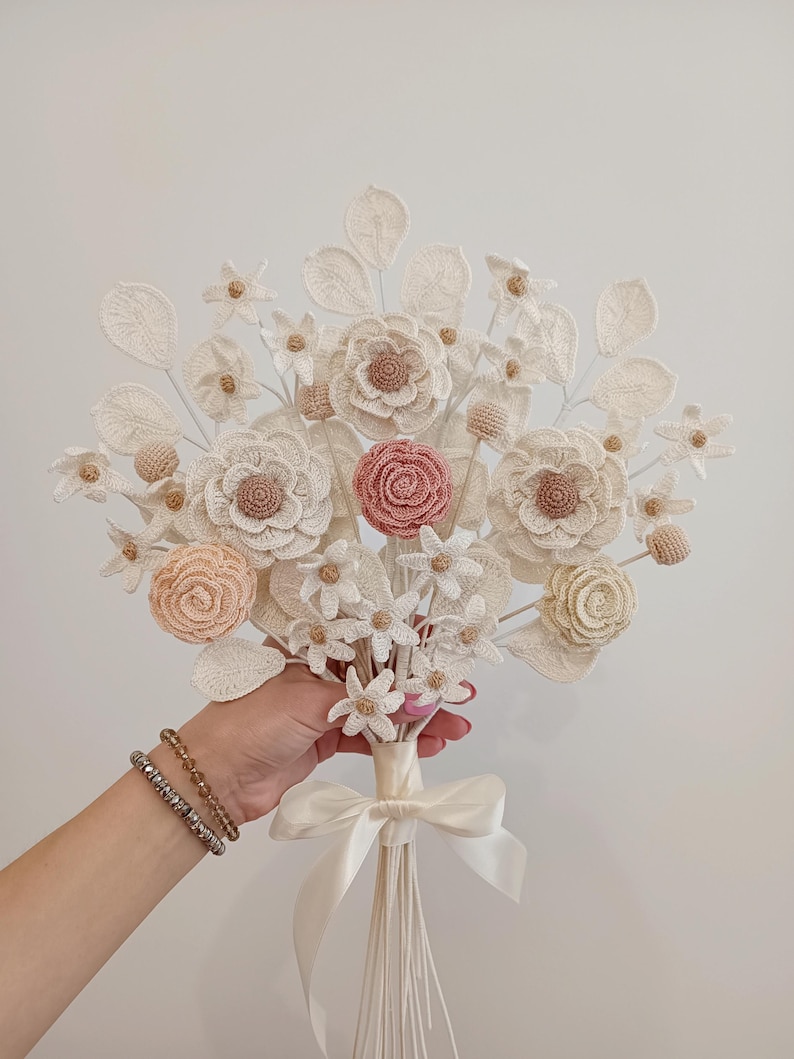 Puede incluir: Un ramo de flores de ganchillo hecho a mano con flores crema, melocot&oacute;n y rosa. El ramo presenta diferentes tipos de flores, detalles de p&eacute;talos y un tallo atado con una cinta. El dise&ntilde;o general es delicado y elegante.