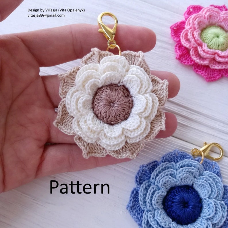 Puede incluir: Tres llaveros de flores de ganchillo en diferentes colores: beige, rosa y azul. Cada flor tiene un anillo de metal dorado. El texto "Pattern" est&aacute; escrito en la superficie blanca debajo de las flores.