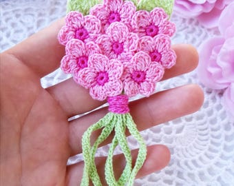 May include: A crochet flower bouquet applique featuring multiple light pink blossoms with bright pink centers, accented by light green leaves at the top and long, thin light green stems tied together with a pink yarn wrap at the base.