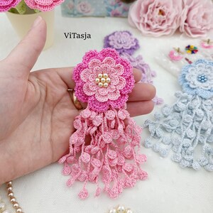 Puede incluir: Un broche de flor de ganchillo rosa con un dise&ntilde;o de p&eacute;talos en capas, un grupo de peque&ntilde;as perlas artificiales de color crema en el centro y un largo fleco de delicados bucles de ganchillo que cuelgan de la base. El texto ViTasja es visible.