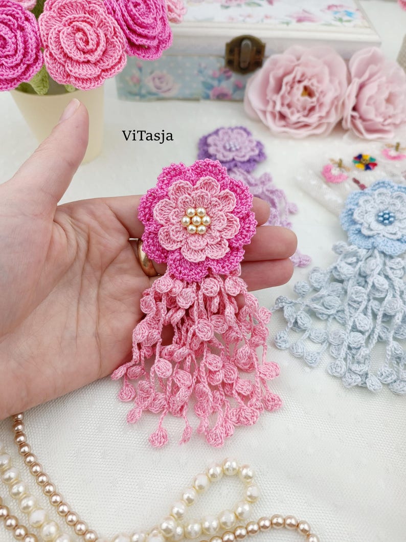 Puede incluir: Un broche de flor de ganchillo rosa con un dise&ntilde;o de p&eacute;talos en capas, un grupo de peque&ntilde;as perlas artificiales de color crema en el centro y un largo fleco de delicados bucles de ganchillo que cuelgan de la base. El texto ViTasja es visible.