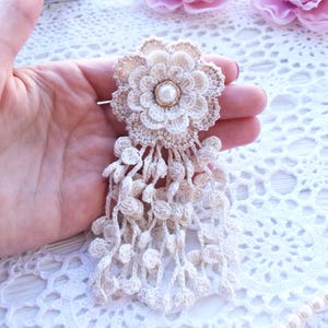 Puede incluir: Un broche de flor de ganchillo beige con una perla artificial blanca central rodeada de p&eacute;talos en capas. La flor est&aacute; unida a un fleco en cascada de delicadas cadenas de ganchillo adornadas con peque&ntilde;as bolas de ganchillo beige.