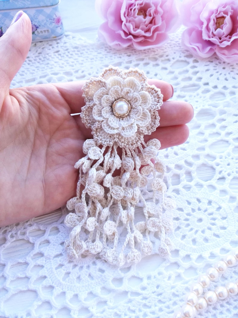 Puede incluir: Un broche de flor de ganchillo beige con una perla artificial blanca central rodeada de p&eacute;talos en capas. La flor est&aacute; unida a un fleco en cascada de delicadas cadenas de ganchillo adornadas con peque&ntilde;as bolas de ganchillo beige.