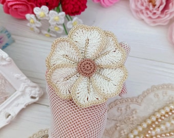May include: A crocheted flower brooch featuring five cream-colored petals with tan edges and a textured, light brown circular center. The flower is crafted with intricate yarn stitching and is displayed against a white fishnet fabric background.
