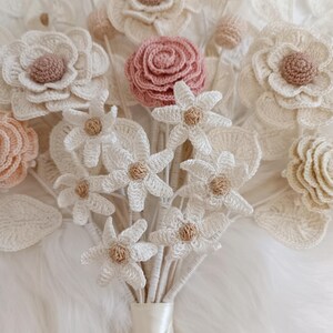 Puede incluir: Un ramo de flores de ganchillo hechas a mano en varios tonos de blanco, crema y rosa rubor. El ramo presenta diferentes tipos de flores, incluyendo rosas y margaritas, con centros beige. Los tallos est&aacute;n atados con una cinta de sat&eacute;n color crema.