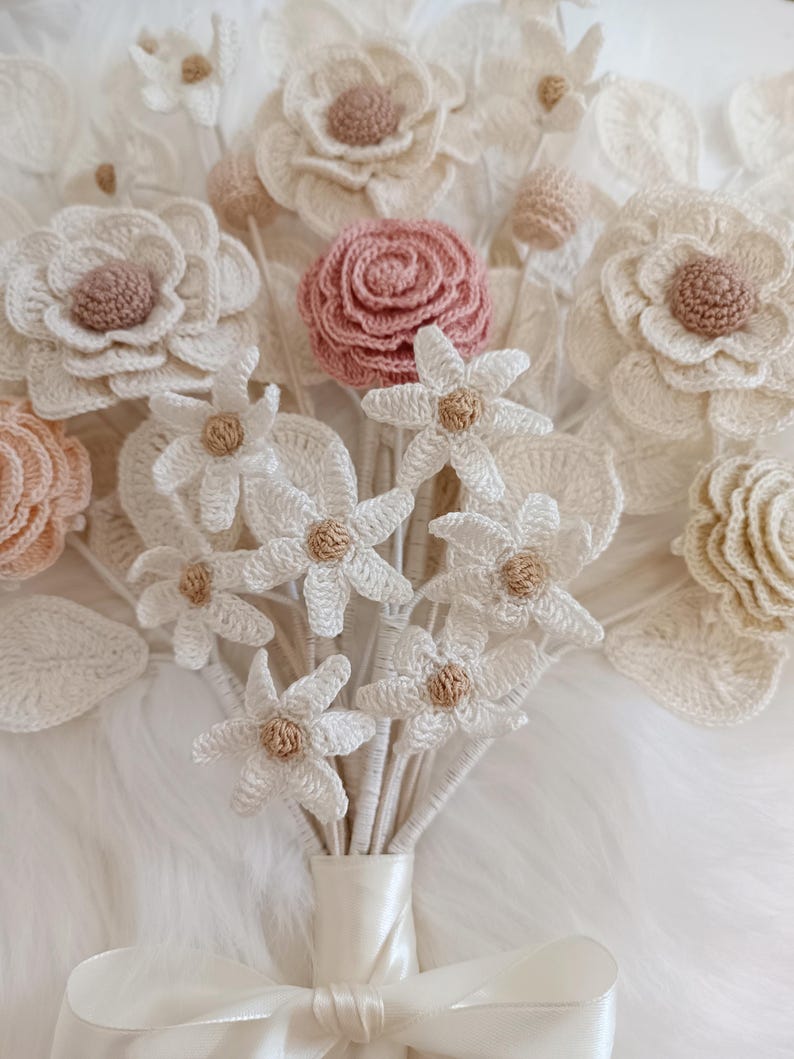 Puede incluir: Un ramo de flores de ganchillo hechas a mano en varios tonos de blanco, crema y rosa rubor. El ramo presenta diferentes tipos de flores, incluyendo rosas y margaritas, con centros beige. Los tallos est&aacute;n atados con una cinta de sat&eacute;n color crema.