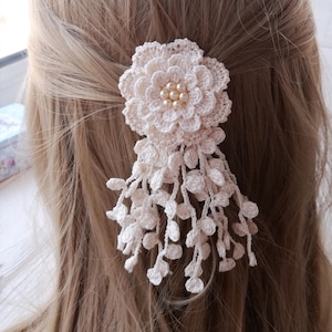 Puede incluir: Un accesorio para el cabello de ganchillo color crema que presenta una flor grande de varias capas con un grupo de peque&ntilde;as perlas redondas de color blanco roto en el centro. Varios delicados hilos de enredadera de ganchillo cuelgan debajo.