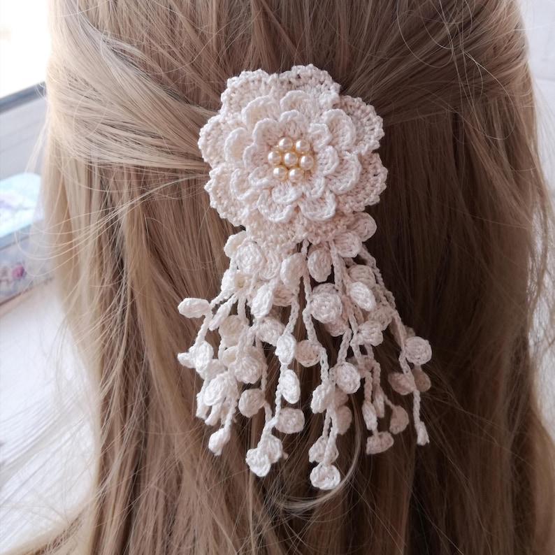 Puede incluir: Un accesorio para el cabello de ganchillo color crema que presenta una flor grande de varias capas con un grupo de peque&ntilde;as perlas redondas de color blanco roto en el centro. Varios delicados hilos de enredadera de ganchillo cuelgan debajo.