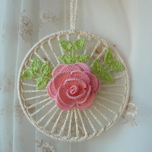 Puede incluir: Un adorno de pared circular tejido a ganchillo con un marco de hilo color crema, una rosa rosa central y hojas de color verde claro. La rosa tiene pétalos superpuestos y las hojas están situadas a ambos lados. Incluye un lazo de hilo a juego en la parte superior para colgar.